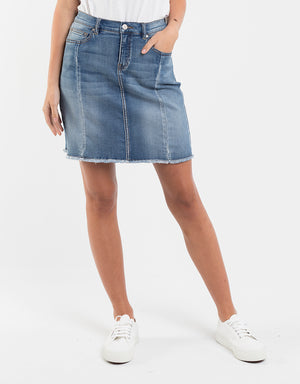 A person in a trendy Foxwood Kiama Skirt with a frayed hem, paired with white sneakers and a white top, stands casually with one hand in their pocket against a plain white background.