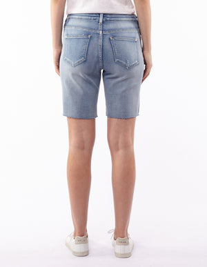 A person is shown from the back wearing the Foxwood AJA Bermuda Short in light blue denim, featuring a frayed hem and distressed detailing. These super soft shorts include back pockets and are paired with a white top and white sneakers. The plain white background highlights the comfortable style.