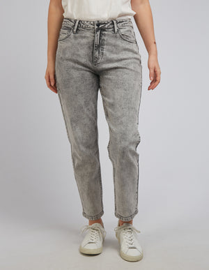 Dressed in white sneakers and a white t-shirt, an individual is posing against a plain gray background in the comfortable Foxwood Barkly Straight Leg Jeans, featuring a subtle patch. The stylish jeans enhance their relaxed stance with arms at their sides.