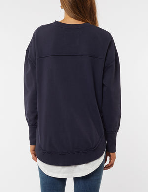 A person with long hair wears a navy blue Foxwood Delilah Crew, featuring raw edge detailing, over a white shirt. The 100% cotton oversized jumper pairs perfectly with blue jeans as they turn away, highlighting the sweatshirt's back.