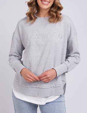 A person smiles while wearing the Foxwood Simplified Crew in light gray, paired with light blue jeans. Their wavy, shoulder-length hair stands out against a plain gray background.