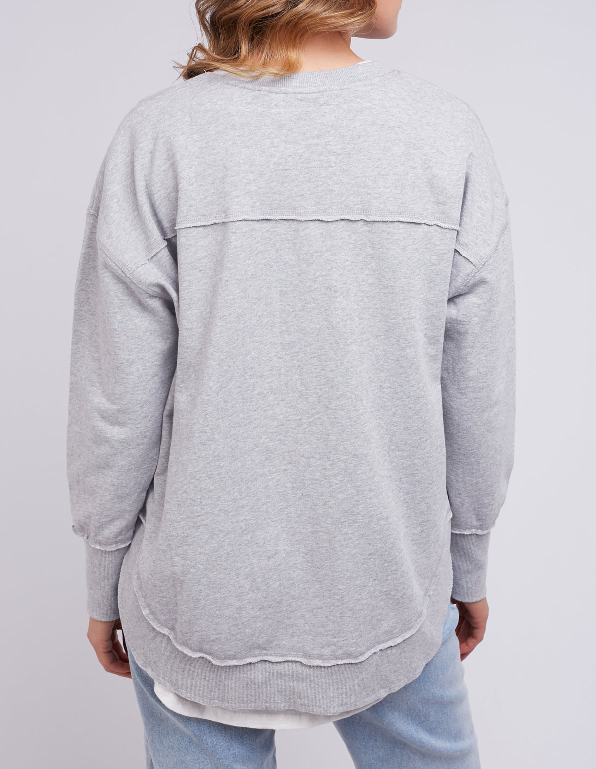 A person wears a gray cotton jumper with seam details and light blue jeans. Facing away from the camera, they showcase the Foxwood Simplified Crew against a plain light gray background.
