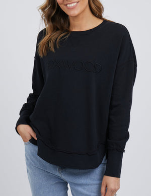 A woman in a relaxed fit black Foxwood Simplified Crew sweatshirt, made from 100% cotton, pairs it effortlessly with light blue jeans. With wavy brown hair cascading over her shoulder, she smiles warmly, one hand casually tucked in her pocket.