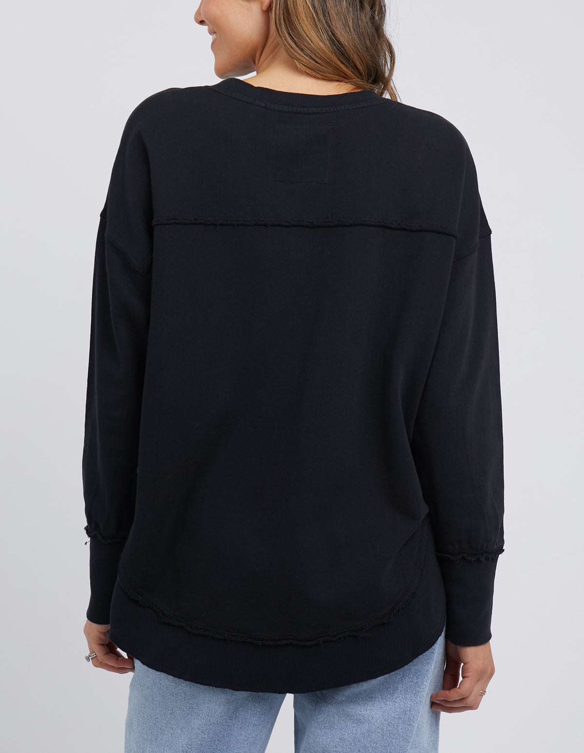 A person in a black, relaxed fit Foxwood Simplified Crew and light blue jeans stands with their back to the camera. The 100% cotton shirt has visible seam details on the upper back and sleeves, adding subtle charm.