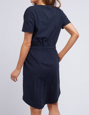 A person is seen from the back wearing a navy Foxwood Ingrid dress, featuring short sleeves and an elastic waist. They stand with one arm slightly bent against a plain light gray background.