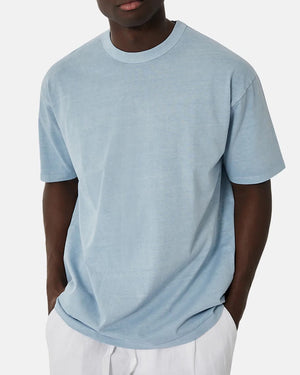 A person wearing the Industrie The Del Sur Tee, a light blue, oversized, comfortable tee, and white pants stands against a plain background with hands in pockets for a relaxed vibe.