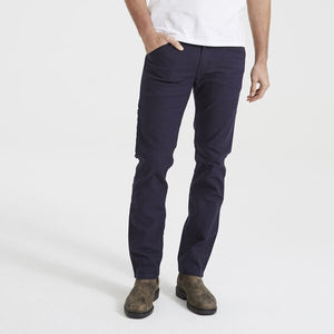 A person wearing dark blue Levis Workwear 511 Utility Pants, a white shirt, and comfortable brown suede boots stands with one hand in their pocket against a plain white background. The image is focused on the pants and shoes, with the head of the person out of the frame.