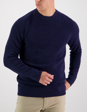 A person poses against a white background wearing the Swanndri Otahu River Crew Neck Merino Knit in navy blue and beige pants. They turn slightly to the side, elegantly adjusting the sweater with one hand.