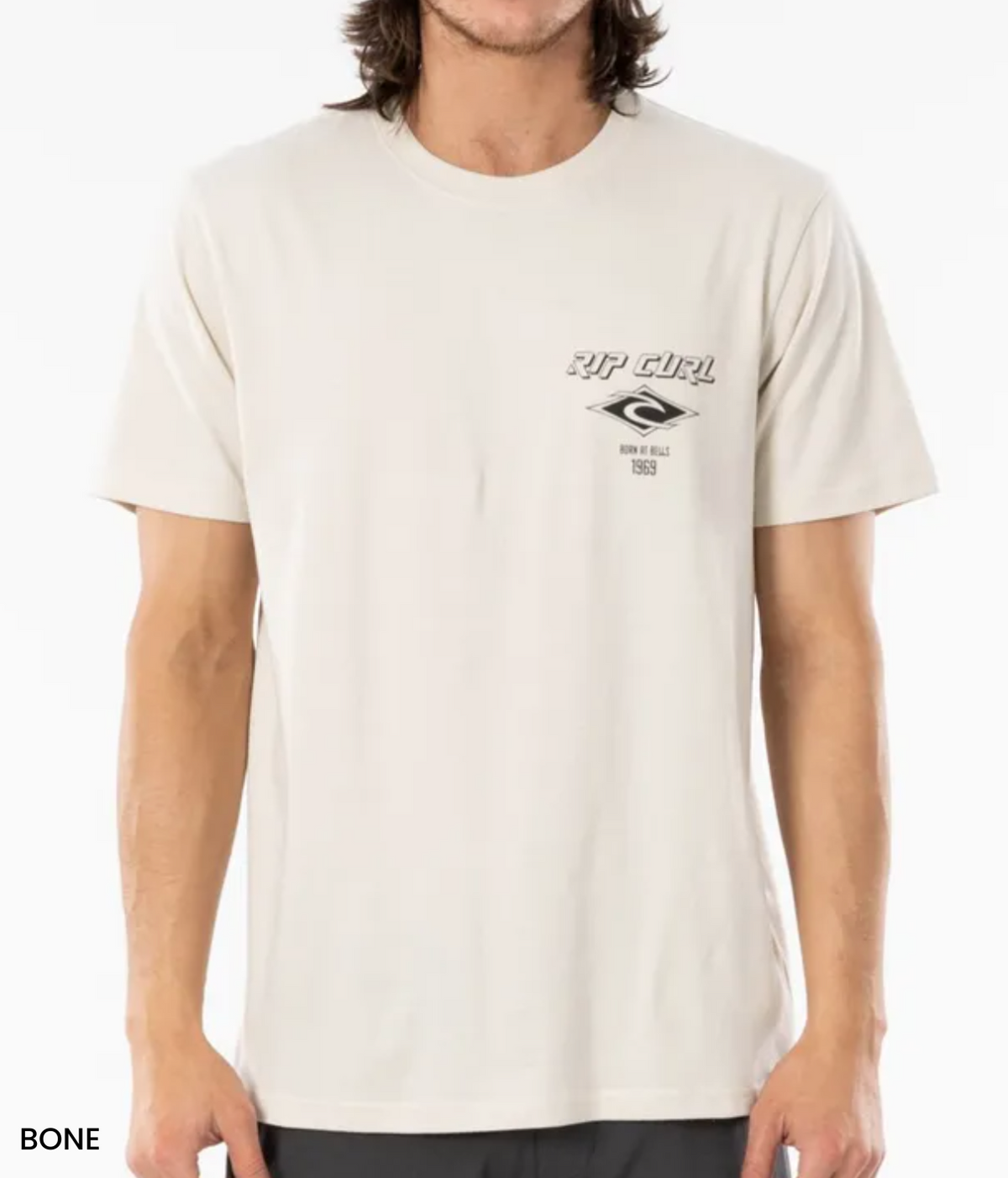 Someone in a light beige Rip Curl Fade Out Icon Tee, crafted from organic cotton with a small logo and text on the upper left, is shown. The tee has a relaxed fit and short sleeves, paired with dark pants against a plain white background.