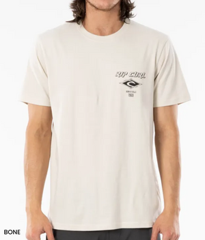 Someone in a light beige Rip Curl Fade Out Icon Tee, crafted from organic cotton with a small logo and text on the upper left, is shown. The tee has a relaxed fit and short sleeves, paired with dark pants against a plain white background.