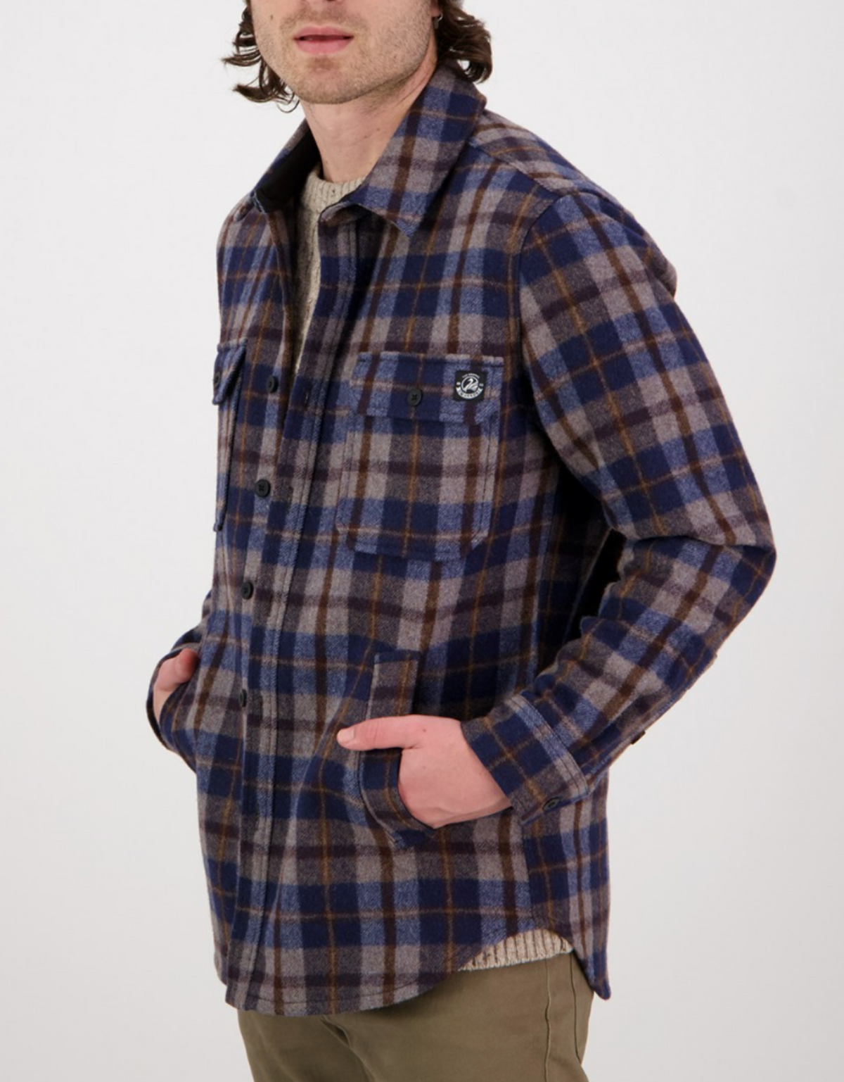 A person wears the Swanndri Kiaraki v3 Wool Shacket in blue, brown, and gray plaid over a beige sweater with olive pants, standing with hands in the shacket’s pockets against a plain white background.