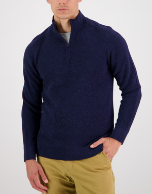 A man wears a navy Swanndri Magnet Bay v2 Merino Jumper over a gray shirt and tan pants, standing with one hand in his pocket against a plain white background.