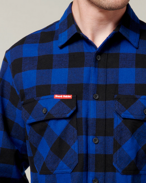 A person wearing a Hard Yakka Check Flannelette Shirt in blue and black checkered cotton twill, featuring front pockets and a distinctive red label on one of them, is visible. The image shows only the person's upper torso and part of their face.