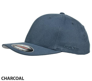 The FlexFit Worn By The World Cap is displayed in a side view, showcasing its stylish and comfortable charcoal gray design. It features a curved contrast brim adorned with a Flexfit sticker and the Flexfit logo embroidered on the side. Set against a plain white background, the word "CHARCOAL" is written in black text at the bottom left corner.