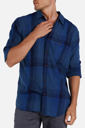 A man wearing the Industrie The Hertford Shirt, a long-sleeve blue plaid button-up with one hand adjusting the collar. The shirt, featuring the distinctive Industrie logo, has a pocket on the left side of the chest. He is also wearing gray pants against a plain white background.
