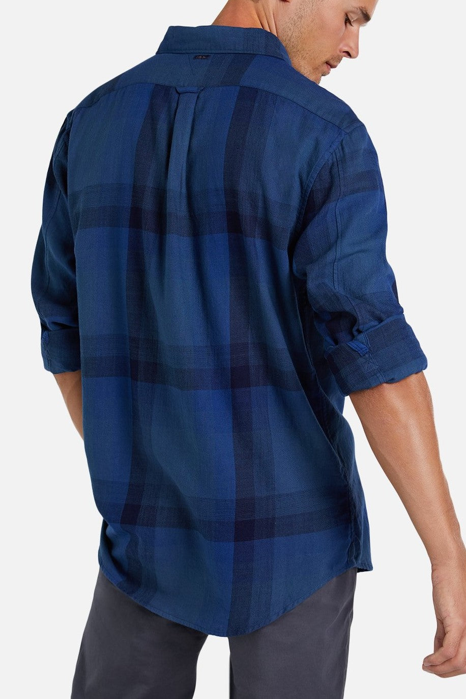 A man is shown from the back wearing Industrie The Hertford Shirt, a blue plaid button-up with rolled-up long sleeves, paired with dark pants. The lightweight shirt features large checkered patterns in various shades of blue and an Industrie logo, standing out against a plain white background.
