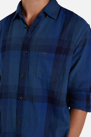 A person sports Industrie The Hertford Shirt, a stylish blue plaid shirt with a collar and an Industrie logo on the chest pocket. The lightweight shirt's long sleeves are rolled up to the elbows, with the image cropped above shoulders and mid-torso.