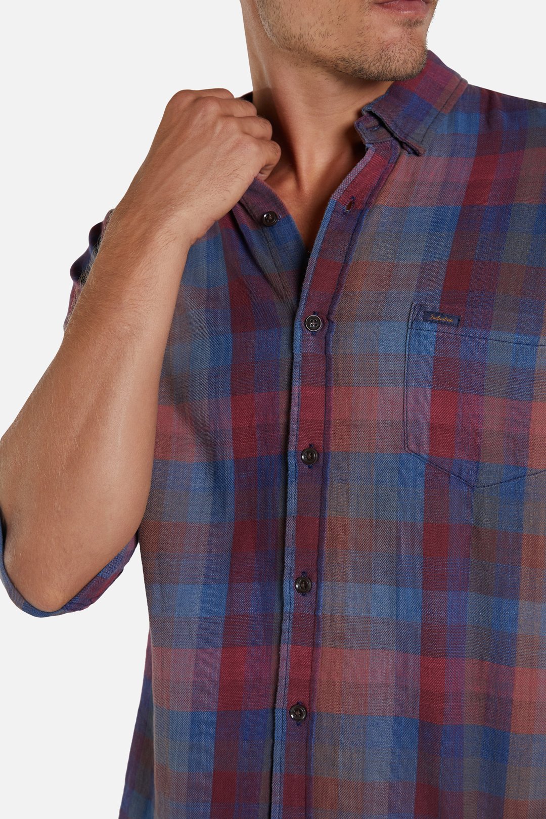 A person wearing an Industrie The Harlow Shirt, featuring a multicolored plaid pattern with rolled-up long sleeves, a button-down front, and a chest pocket, is shown. Their face is partially visible as they adjust the collar with one hand against a plain white background.