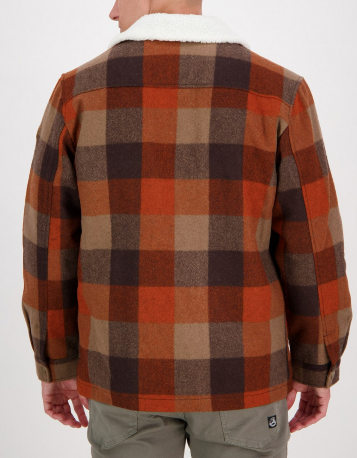 A person wearing the Swanndri Kaituna Sherpa Lined Jacket in brown, orange, and beige plaid with a cozy sherpa collar is seen from the back, paired with light gray pants.
