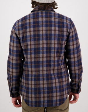A person stands with their back to the camera, wearing a blue and brown Swanndri Kiaraki v3 Wool Shacket, olive green pants, and a light gray beanie against a plain white background.