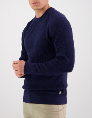A person wearing the Swanndri Otahu River Crew Neck Merino Knit in navy and beige pants stands against a plain white background, with their face turned to the side and not visible.