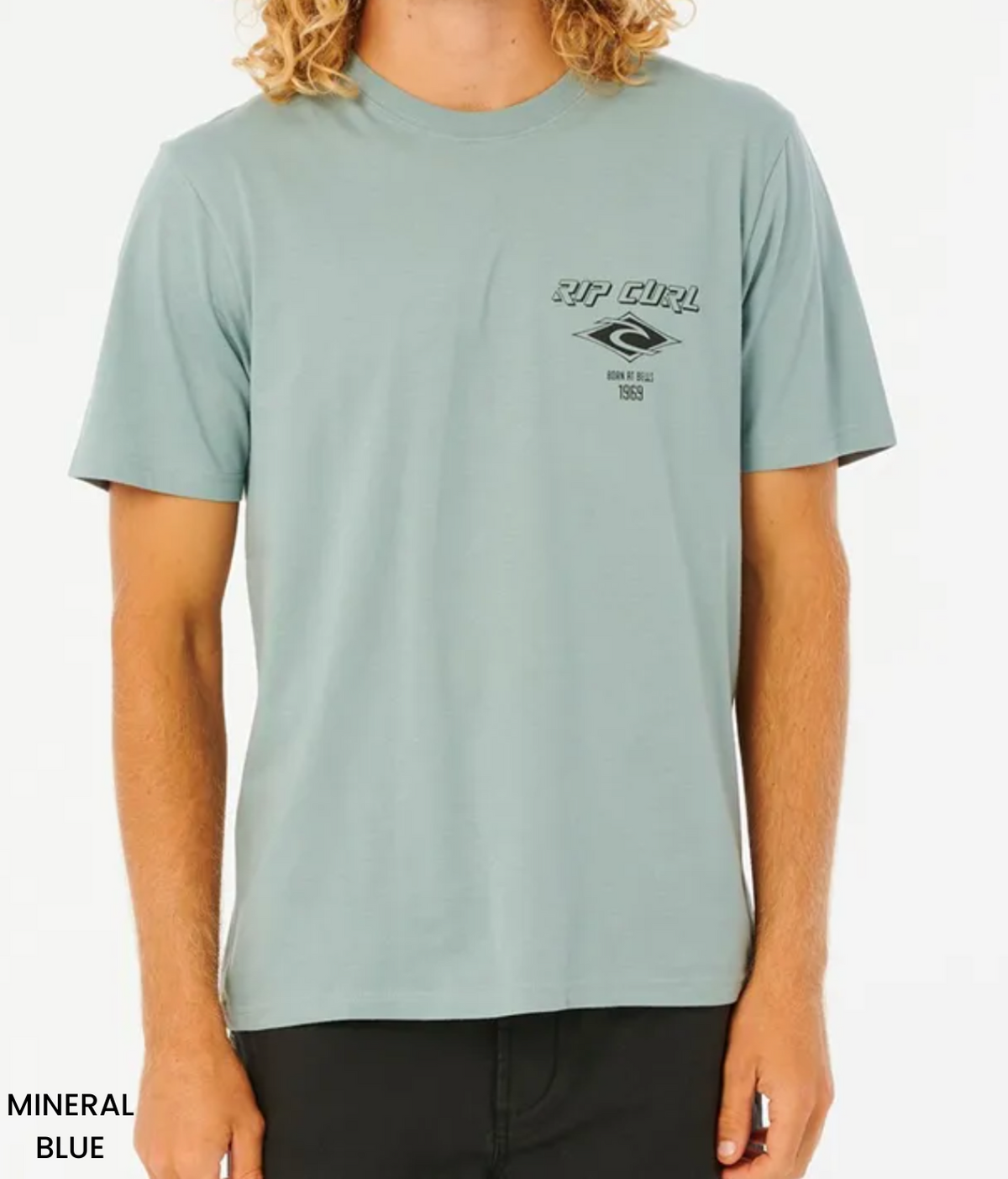 Wearing a mineral blue Rip Curl Fade Out Icon Tee made from organic cotton, displaying "RIP CURL" and "Born at Bells 1969," the person with shoulder-length, wavy blonde hair completes their look with relaxed-fit black pants.