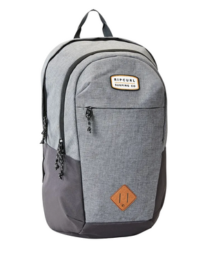 The Rip Curl Overtime 30L Driven Backpack is a gray and black model crafted from recycled polyester. It features a brown "Ripcurl Surfing Co" patch on the top front pocket and includes multiple zippered compartments, a handle, laptop compatibility, and eco-friendly materials.