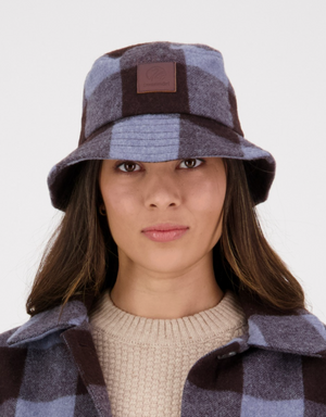 A person wears a Swanndri Kaimai Wool Bucket Hat in blue and brown check, paired with a matching jacket and beige merino sweater, looking at the camera. The hat has a leather patch logo on the front against a plain white background.