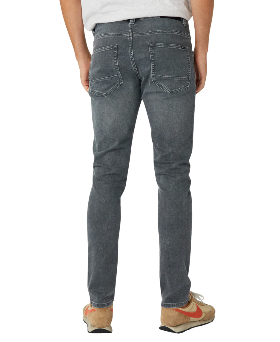 A person in Industrie The Denim Drifter NC Pant, gray denim jeans, and a light gray T-shirt is seen from behind. They are wearing brown and orange sneakers while standing casually against a white backdrop, highlighting the sleek skinny fit.