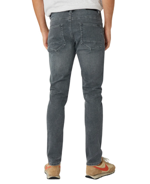 A person in Industrie The Denim Drifter NC Pant, gray denim jeans, and a light gray T-shirt is seen from behind. They are wearing brown and orange sneakers while standing casually against a white backdrop, highlighting the sleek skinny fit.