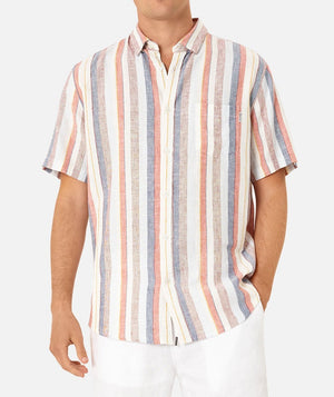 A person is wearing the Industrie The Delgrado Short Sleeve Shirt, a stylish short-sleeved button-up with vertical stripes in red, beige, white, and blue colors. The classic fit shirt features a front pocket on the left side. The individual has paired this comfortable shirt with white pants and has their left hand in their pocket.