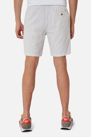 A person is seen from the rear wearing Industrie The Elio Short, made of light gray linen-cotton. It has an elasticized waist, a single back pocket, and white vertical stripes. They're paired with Nike sneakers that feature red and white details against a plain white background.