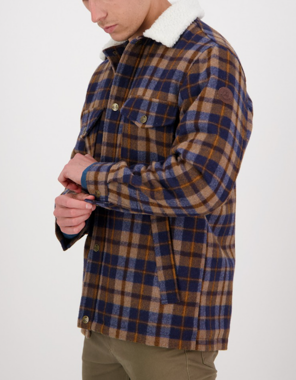 A man in olive green pants buttons the cuff of his Swanndri Kaituna Sherpa Lined Jacket—a brown, blue, and beige plaid piece with a white sherpa collar and cozy sherpa lining. Only the lower part of his face is visible.