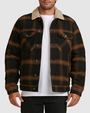 A man stands against a light grey background wearing the Billabong Barlow Sherpa Jacket with a tan sherpa collar, paired with a white t-shirt and black pants.