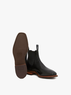 The RM Williams Comfort Kangaroo Craftsman Boot, a pair of black leather Chelsea boots handcrafted in Australia with brown leather soles, is displayed. One boot stands upright while the other is tilted to showcase its brown sole. Both elastic-sided boots feature black pull tabs with white detailing at the back, set against a plain white background.