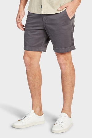 A person wearing a beige shirt paired with dark gray Academy Brand Cooper Chino Shorts and white sneakers. The person has their hands in their pockets and is standing with one leg slightly in front of the other against a plain white background.