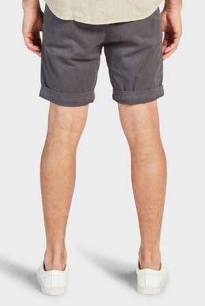 Rear view of a person wearing the dark gray Academy Brand Cooper Chino Shorts, rolled up at the hem, paired with a beige shirt and white sneakers. The person stands with their legs slightly apart against a plain, light-colored background.