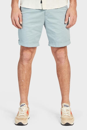 A person stands against a plain white background, wearing Academy Brand Cooper Chino Shorts in light blue cotton that are rolled up at the hem and a light-colored shirt. The person is also sporting beige and white sneakers with hints of orange. Only the person's legs and lower torso are visible.
