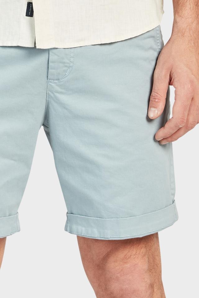 A person wearing Academy Brand Cooper Chino Shorts in light blue, paired with a white shirt. Only the lower torso and upper leg area of the person is visible. The person's right hand is resting naturally by their side. The background is plain and light-colored.