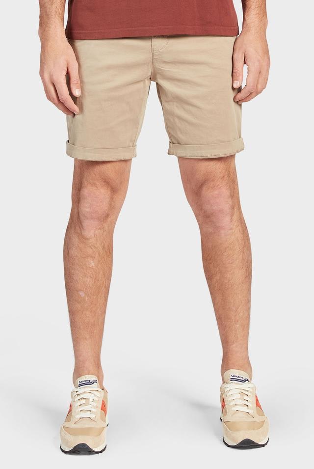 A person is standing against a neutral background, wearing a pair of Academy Brand Cooper Chino Shorts in beige and a maroon shirt. Their outfit is completed with beige and white sneakers that have red and blue accents. Only the lower half of the person's body is visible.