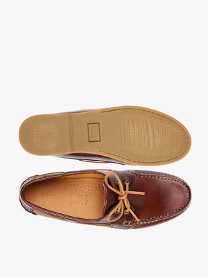 The RM Williams Hobart Boat Shoe features brown yearling leather with tan laces and stitching. A lace bow tops the view, and a light brown rubber outsole is visible on one shoe turned upside down to show the sole, embodying elegant boat shoe style.