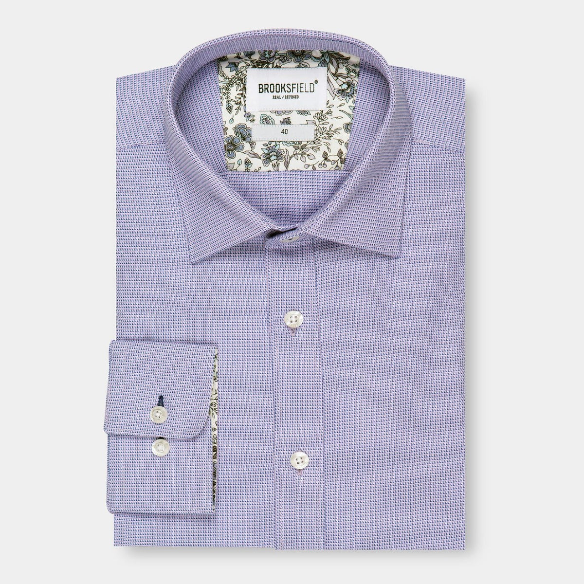 A Brooksfield Micro Textured Shirt, neatly folded and featuring a light purple hue with long sleeves. This stylish shirt showcases a small check pattern along with delicate floral inner collar and cuff linings, all elegantly displayed against a white background.