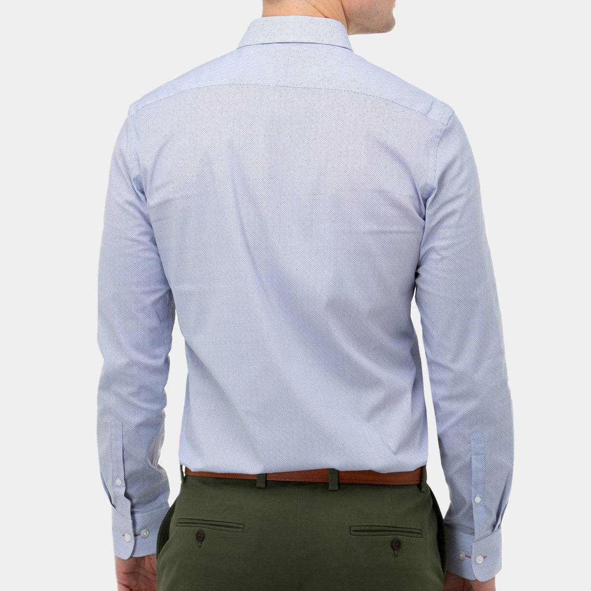 A person is shown from the back, wearing a light blue Brooksfield Stretch Print Shirt paired with green regular fit trousers. The shirt features a stiff point collar and buttoned cuffs, and it is tucked into the trousers. The person is also sporting a brown belt against a white background.