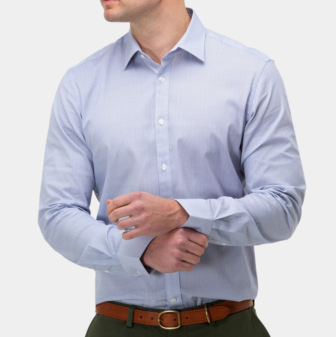 A person wearing a Brooksfield Stretch Print Shirt in light blue with a point collar is adjusting their cuff. They have dark green pants and a brown belt. The background is plain light gray, highlighting the formal attire. The person's face is not fully visible in the image.