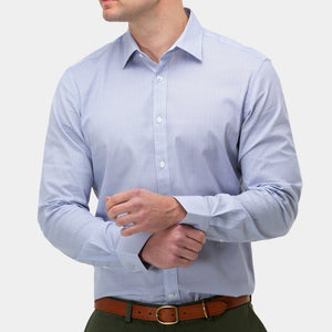 A person wearing a Brooksfield Stretch Print Shirt in light blue with a point collar is adjusting their cuff. They have dark green pants and a brown belt. The background is plain light gray, highlighting the formal attire. The person's face is not fully visible in the image.