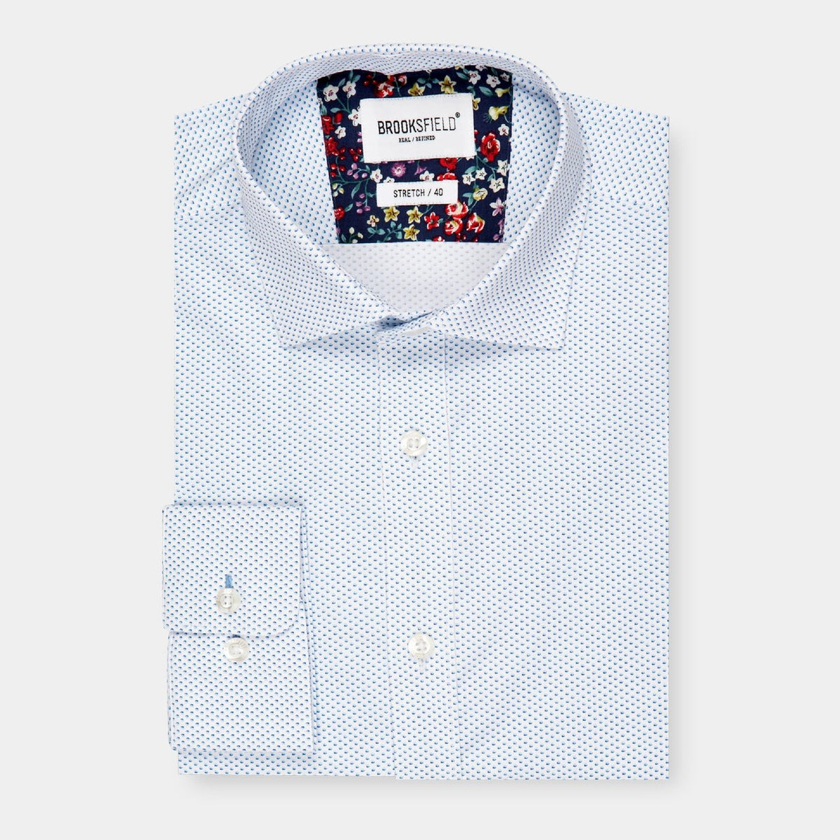 A neatly folded, stylish Brooksfield Stretch Print Shirt features a white button-up design adorned with small blue polka dots. The inner collar boasts a colorful floral pattern, adding a touch of flair. The label reads "Brooksfield, Stretch, Slim Fit." This comfortable shirt is displayed against a pristine white background.