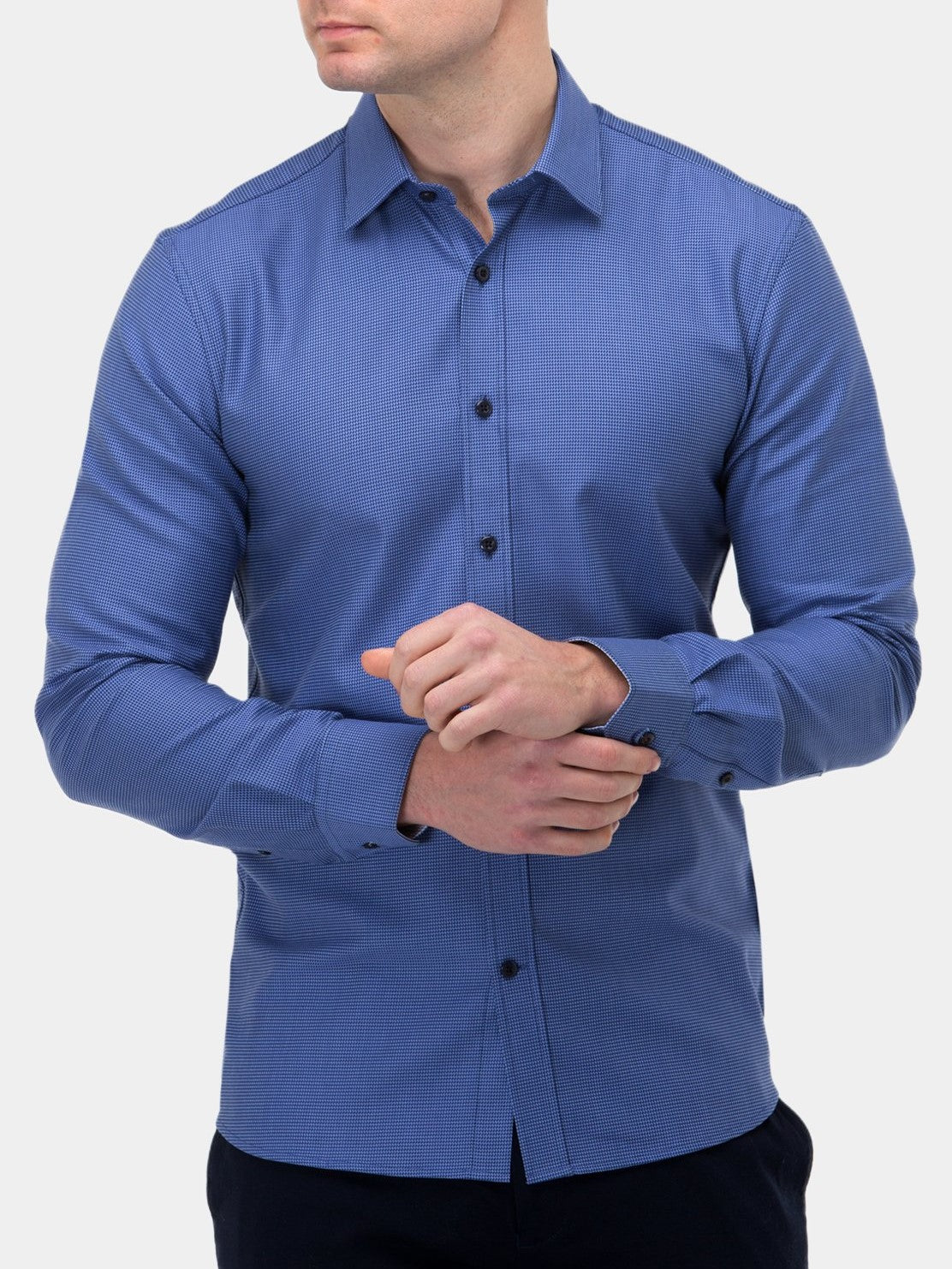A man is wearing a stylish Brooksfield Luxe Textured Shirt, made of 100% cotton in blue with black buttons. He pairs it with dark pants and is seen adjusting the cuff of his left sleeve with his right hand against a plain white background.