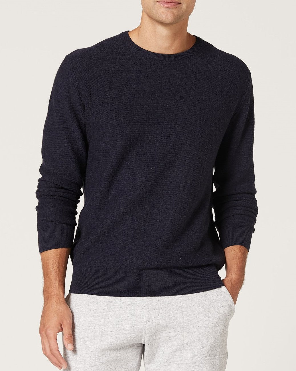 A man wearing a navy Blazer Mason Cotton Crew Neck jumper and light gray pants stands with one hand in his pocket against a plain light background.