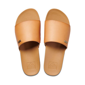 The REEF Cushion Scout slides are crafted from tan leather and feature flat soles. Each sandal has a wide strap across the top adorned with a small metallic label displaying the brand name "REEF." The insole, embossed with the brand's logo, incorporates a cushion bounce midsole and an arch support heel cup for enhanced comfort.