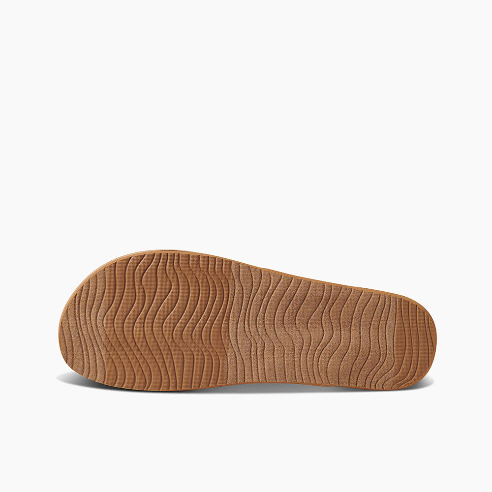 The image shows the bottom view of a REEF Cushion Strand sandal with a light brown sole. The sole features a wavy, ripple-like pattern providing texture and grip. Enhanced arch support and a cushion bounce midsole are highlighted against the plain white background, emphasizing the unique tread design of the shoe sole.